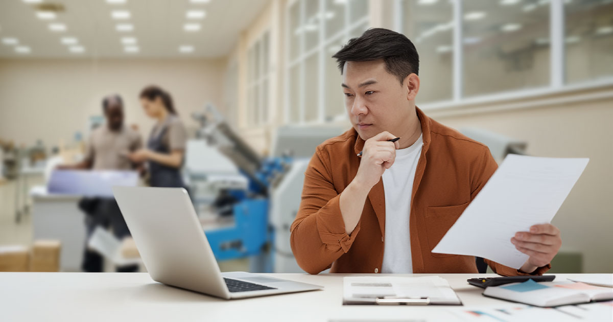 person at a desk reviewing documents, laptop open, looking considered